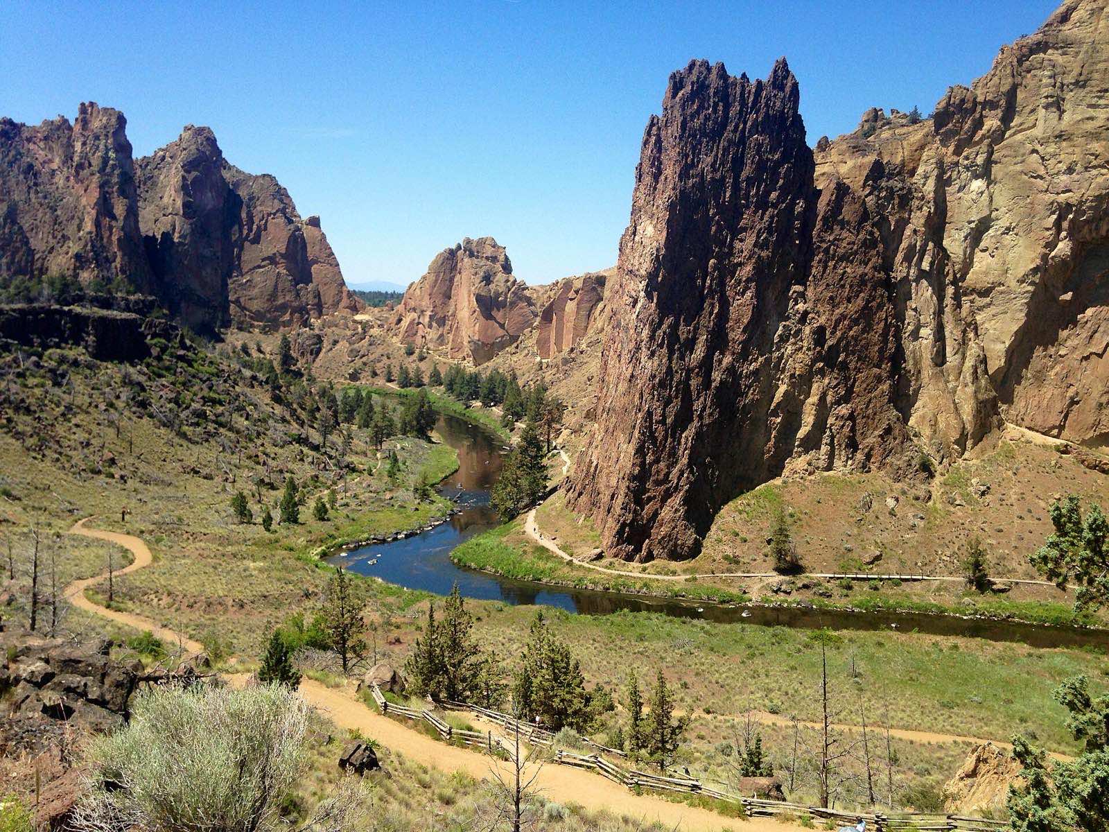 Smith Rock's volcanic spires rising above a bend in the Crooked River, with a trail winding along the canyon floor, Deschutes County, Oregon.