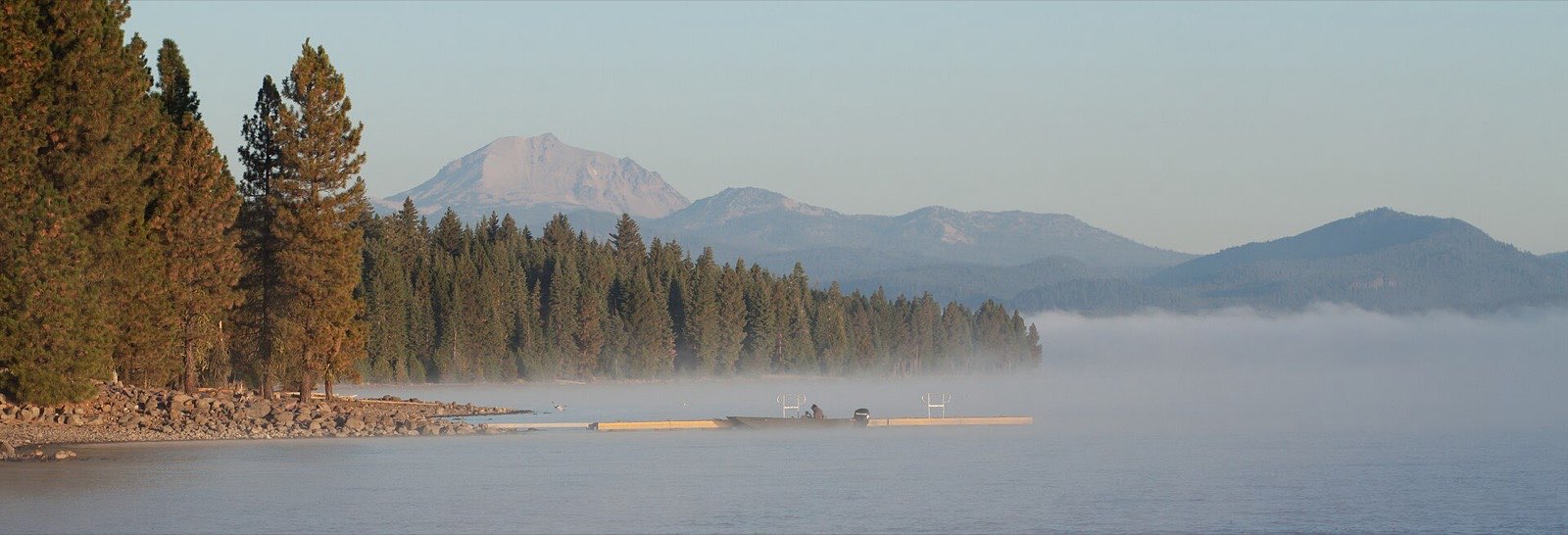 Morning fog rising off Lake Almanor's western shore, framed by pine trees, in Plumas County, California.