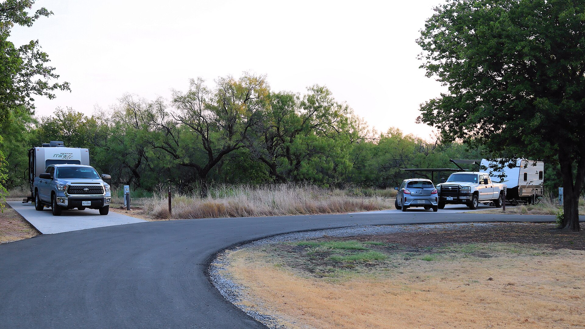 Recreational vehicles and trucks parked at paved sites along a wooded campground loop at dusk.